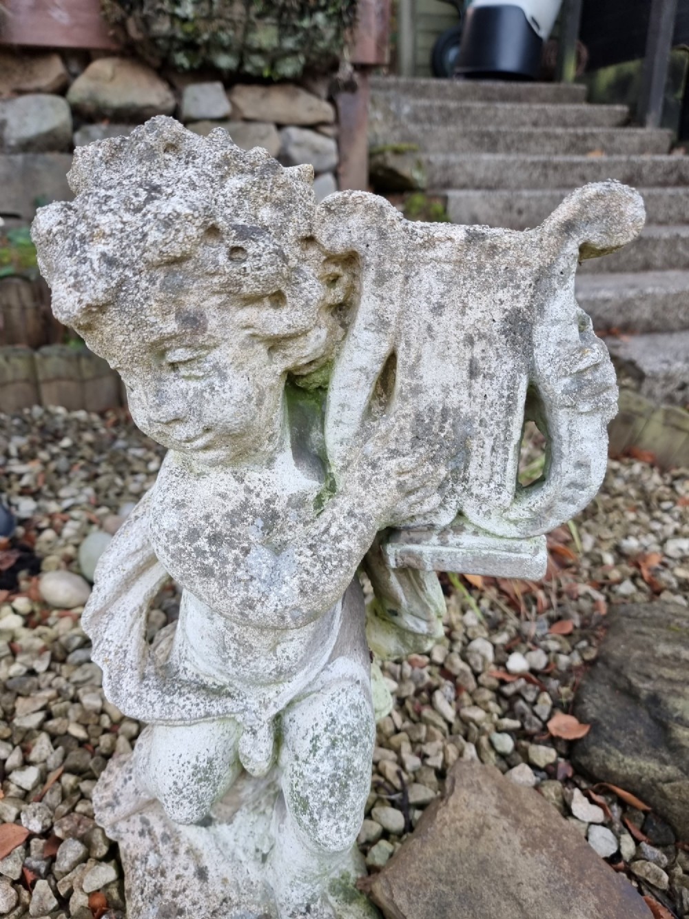 A pair of weathered grey reconstituted stone garden statues depicting classical musical cherubs holding instruments. View IV