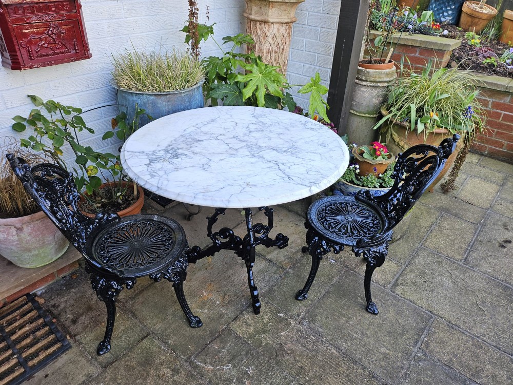 A three-piece bistro set featuring a circular white marble tabletop on a black cast-iron tripod base, accompanied by two matching ornate cast-iron chairs. View II