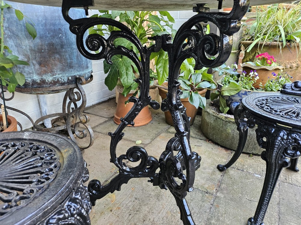 A three-piece bistro set featuring a circular white marble tabletop on a black cast-iron tripod base, accompanied by two matching ornate cast-iron chairs. View III