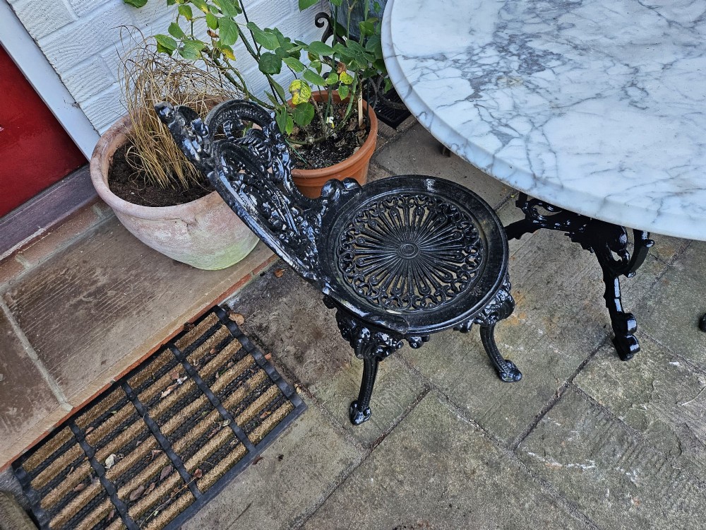 A three-piece bistro set featuring a circular white marble tabletop on a black cast-iron tripod base, accompanied by two matching ornate cast-iron chairs. View IV