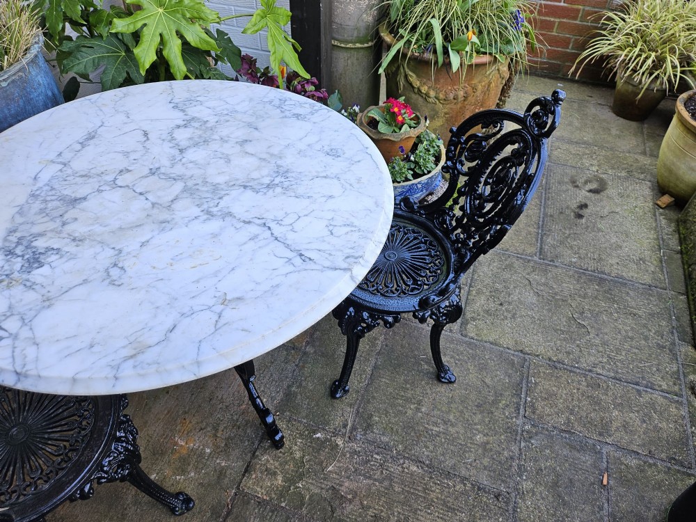 A three-piece bistro set featuring a circular white marble tabletop on a black cast-iron tripod base, accompanied by two matching ornate cast-iron chairs. View V
