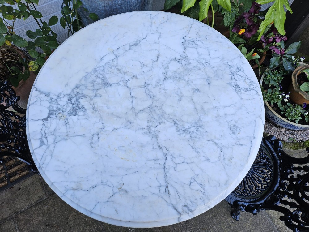 A three-piece bistro set featuring a circular white marble tabletop on a black cast-iron tripod base, accompanied by two matching ornate cast-iron chairs. View VI