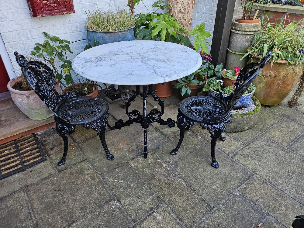 A three-piece bistro set featuring a circular white marble tabletop on a black cast-iron tripod base, accompanied by two matching ornate cast-iron chairs. View VII