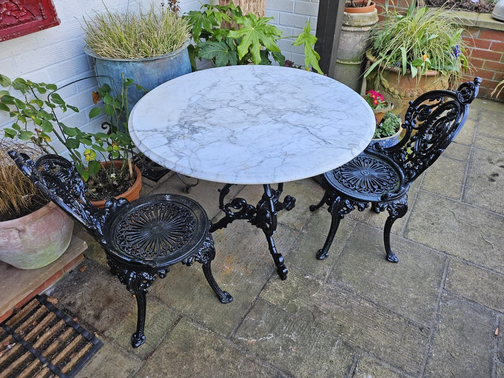 A three-piece bistro set featuring a circular white marble tabletop on a black cast-iron tripod base, accompanied by two matching ornate cast-iron chairs. View VIII