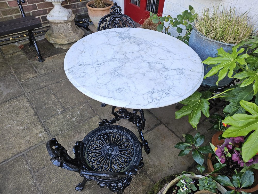 A three-piece bistro set featuring a circular white marble tabletop on a black cast-iron tripod base, accompanied by two matching ornate cast-iron chairs. View IX