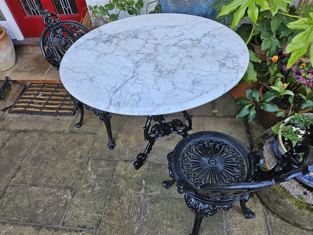 A three-piece bistro set featuring a circular white marble tabletop on a black cast-iron tripod base, accompanied by two matching ornate cast-iron chairs. View X