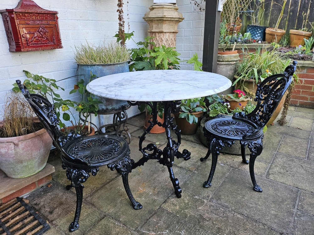 A three-piece bistro set featuring a circular white marble tabletop on a black cast-iron tripod base, accompanied by two matching ornate cast-iron chairs. View XI