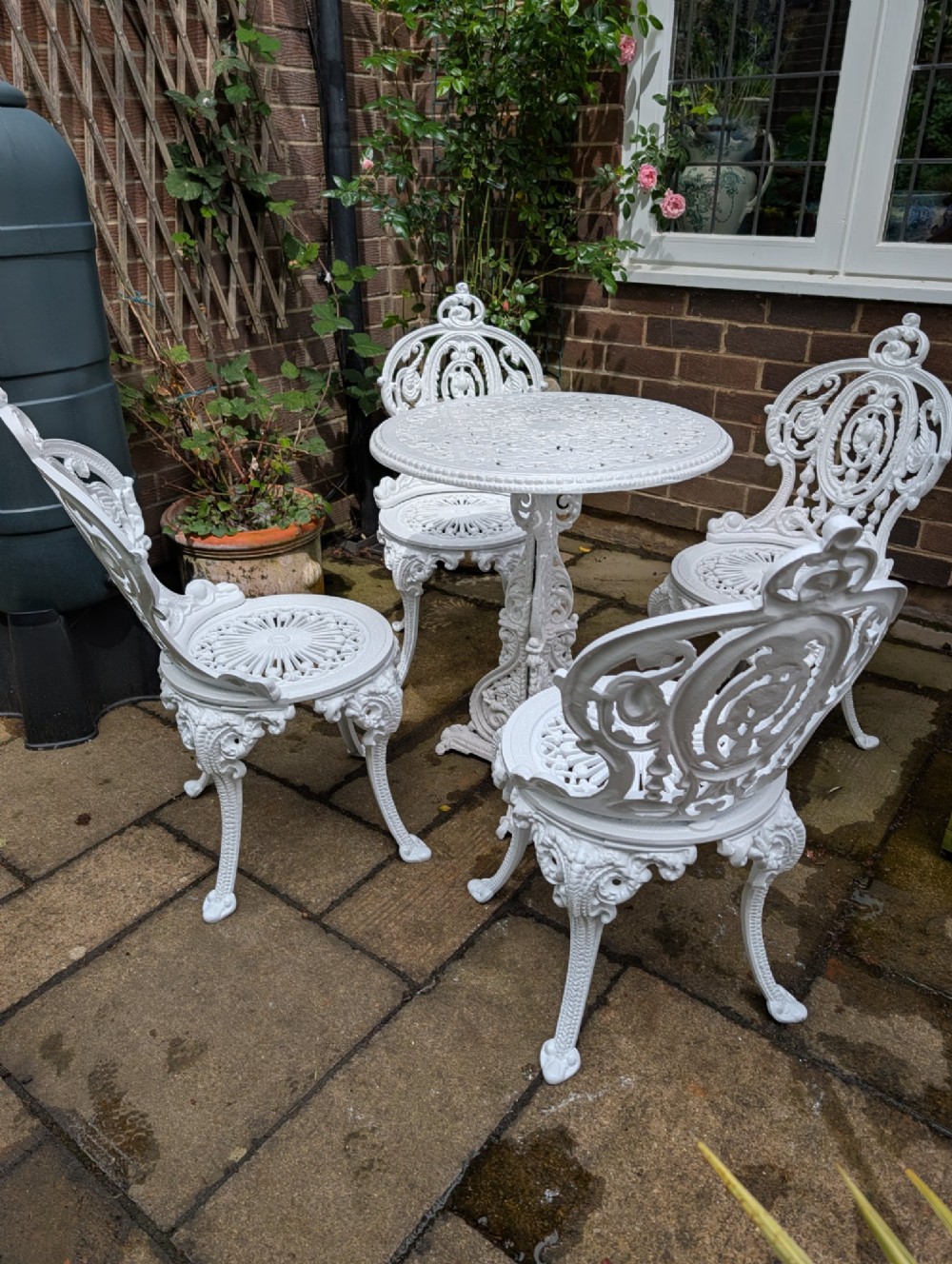 A white painted cast-iron garden set consisting of a round pedestal table and four ornate openwork chairs on a stone patio.