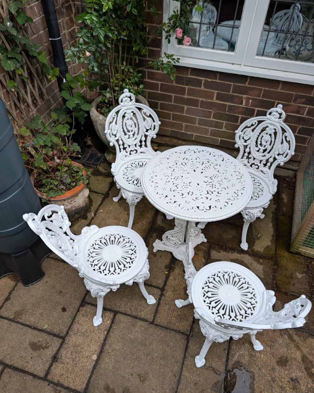 A white painted cast-iron garden set consisting of a round pedestal table and four ornate openwork chairs on a stone patio. View II