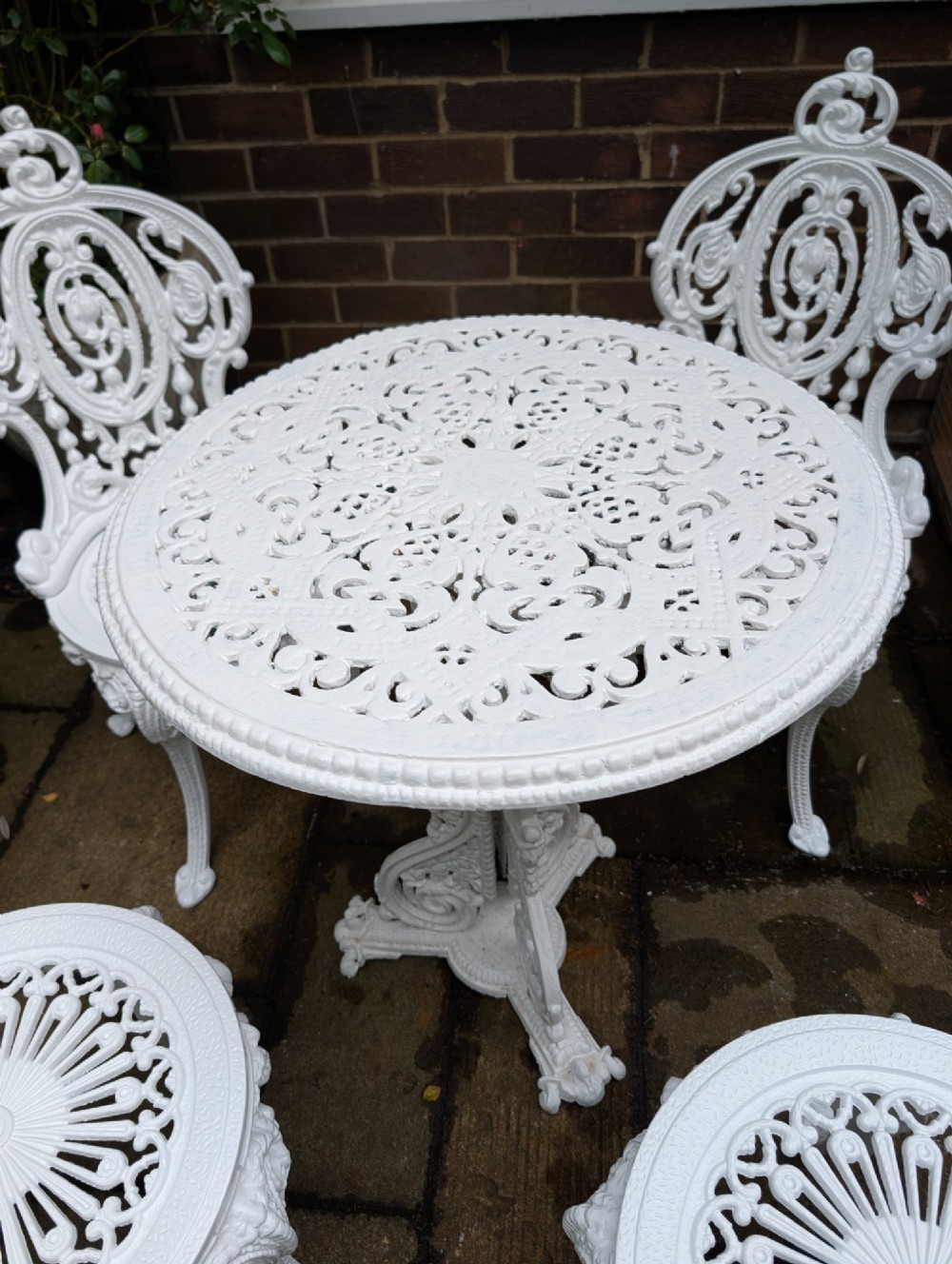 A white painted cast-iron garden set consisting of a round pedestal table and four ornate openwork chairs on a stone patio. View III