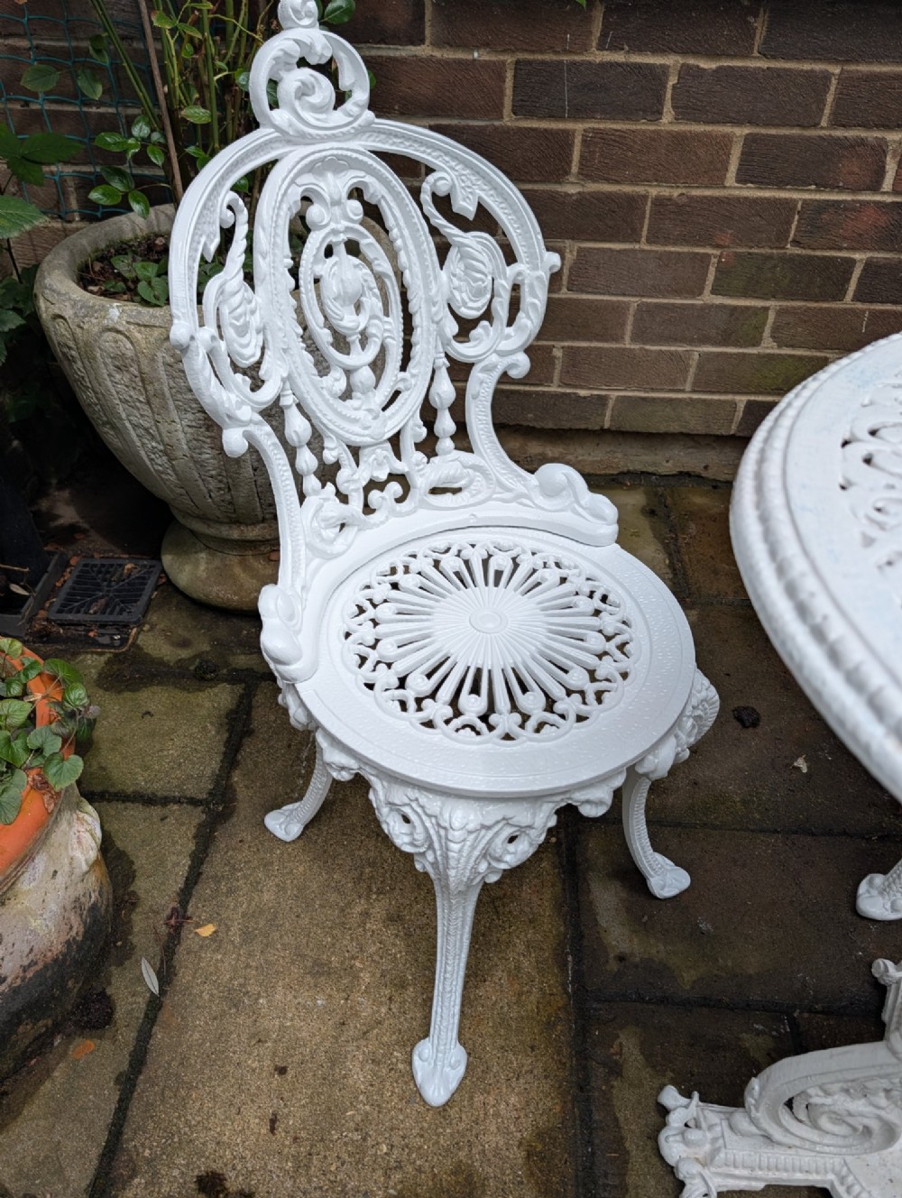 A white painted cast-iron garden set consisting of a round pedestal table and four ornate openwork chairs on a stone patio. View V