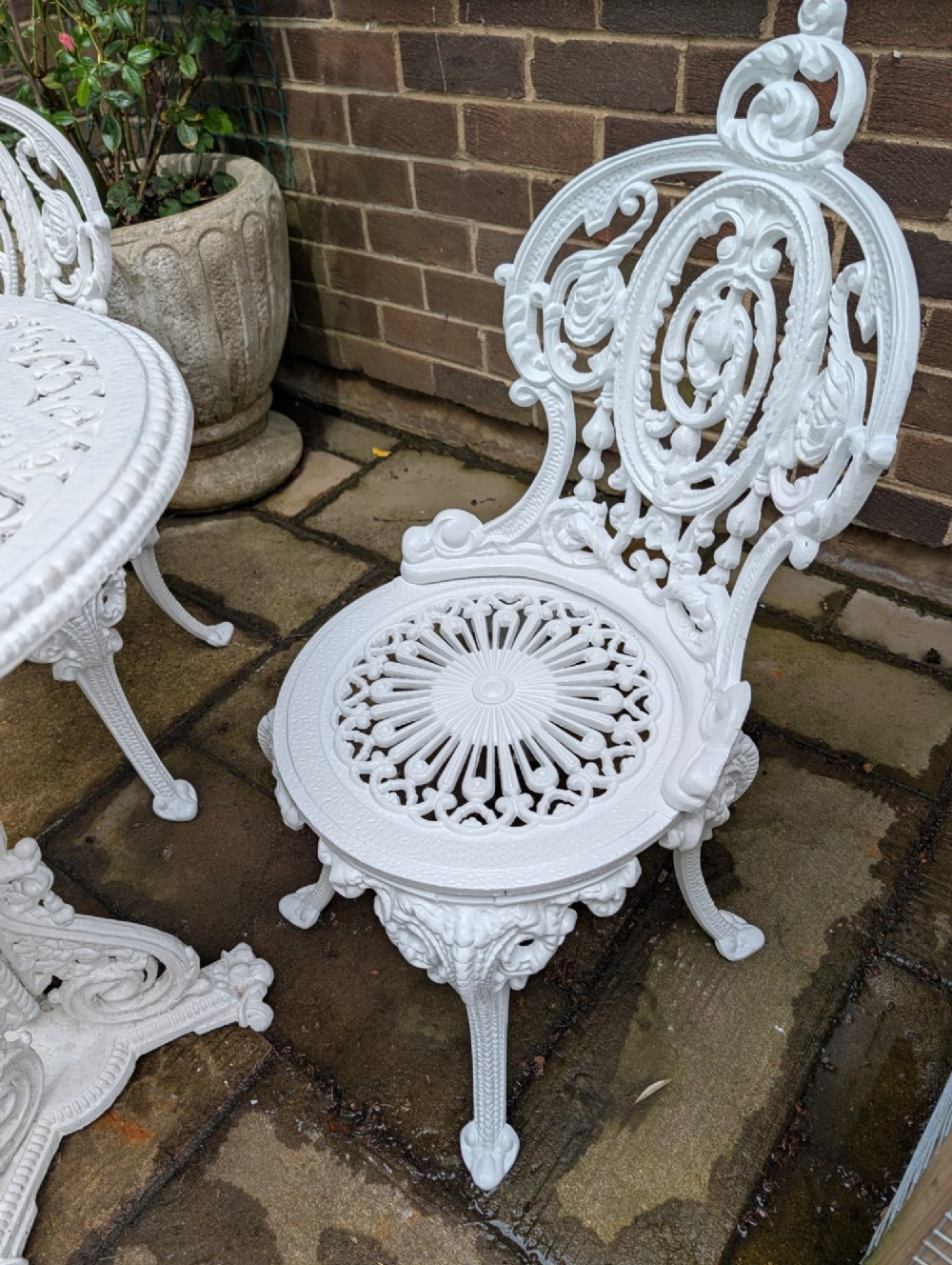 A white painted cast-iron garden set consisting of a round pedestal table and four ornate openwork chairs on a stone patio. View VI
