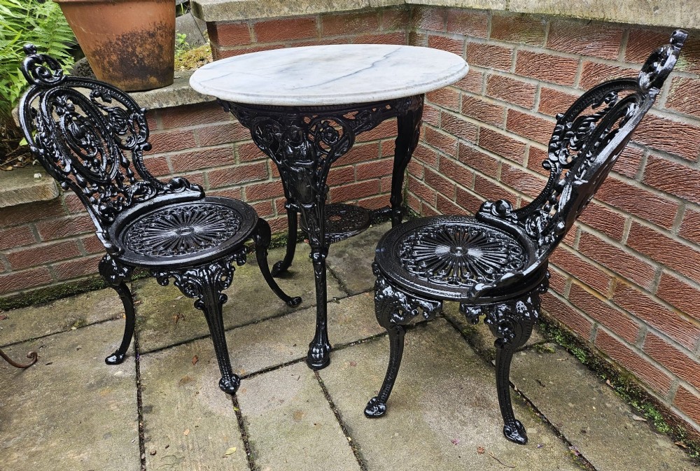 A circular white marble-topped table with a black cast-iron base alongside two matching black cast-iron chairs featuring ornate pierced scrollwork.