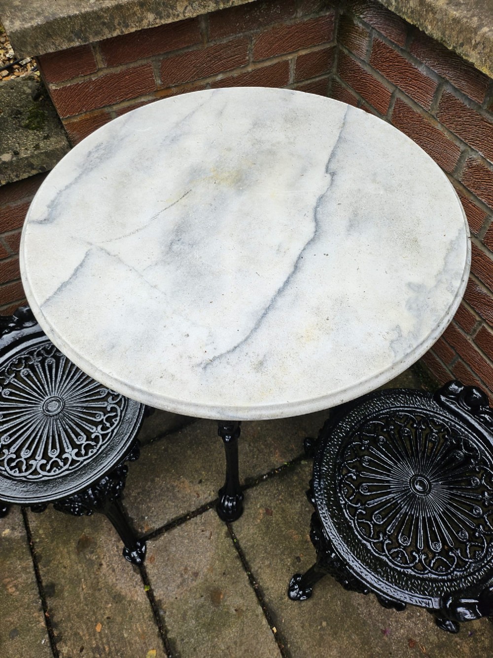 A circular white marble-topped table with a black cast-iron base alongside two matching black cast-iron chairs featuring ornate pierced scrollwork. View VI