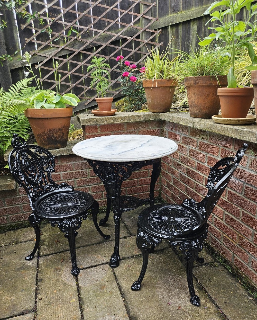 A circular white marble-topped table with a black cast-iron base alongside two matching black cast-iron chairs featuring ornate pierced scrollwork. View X