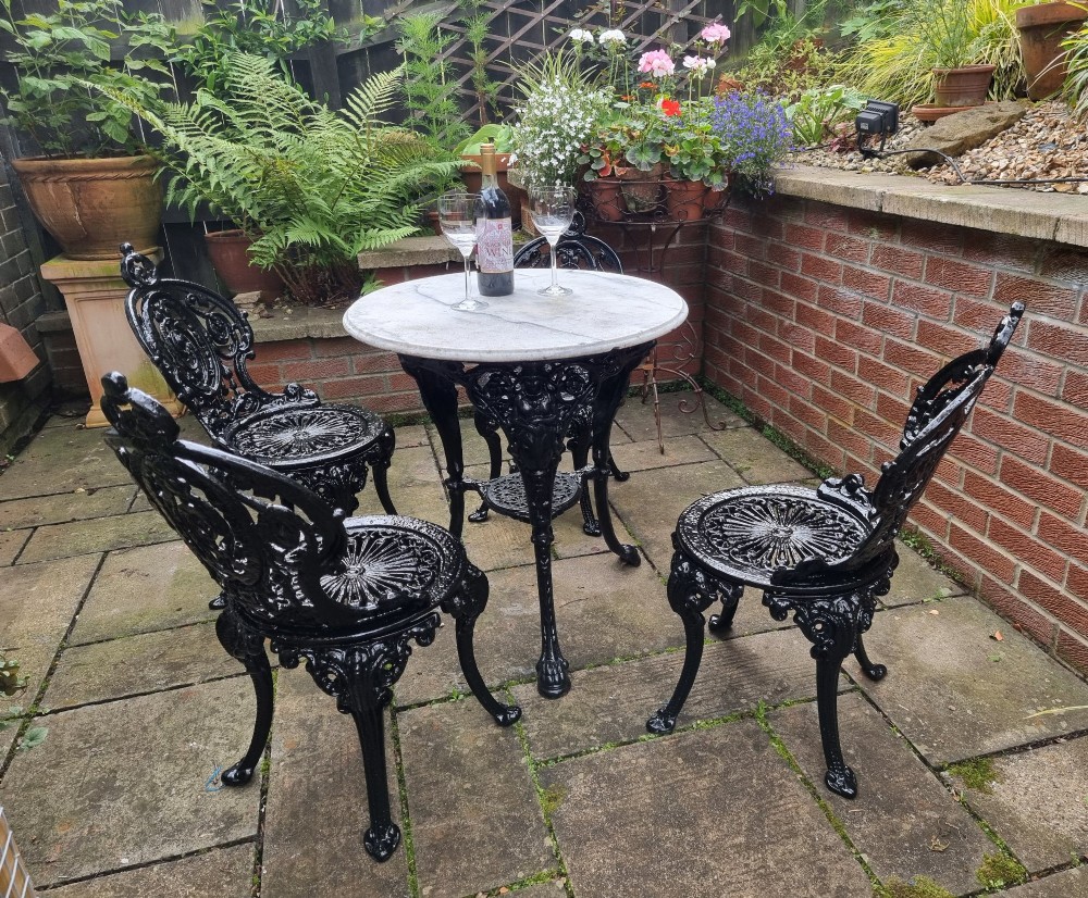 A five-piece garden set featuring a circular white marble-topped table and four black cast iron chairs with ornate pierced floral backrests. View II