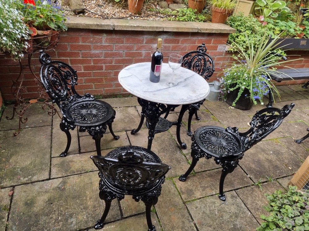 A five-piece garden set featuring a circular white marble-topped table and four black cast iron chairs with ornate pierced floral backrests. View III