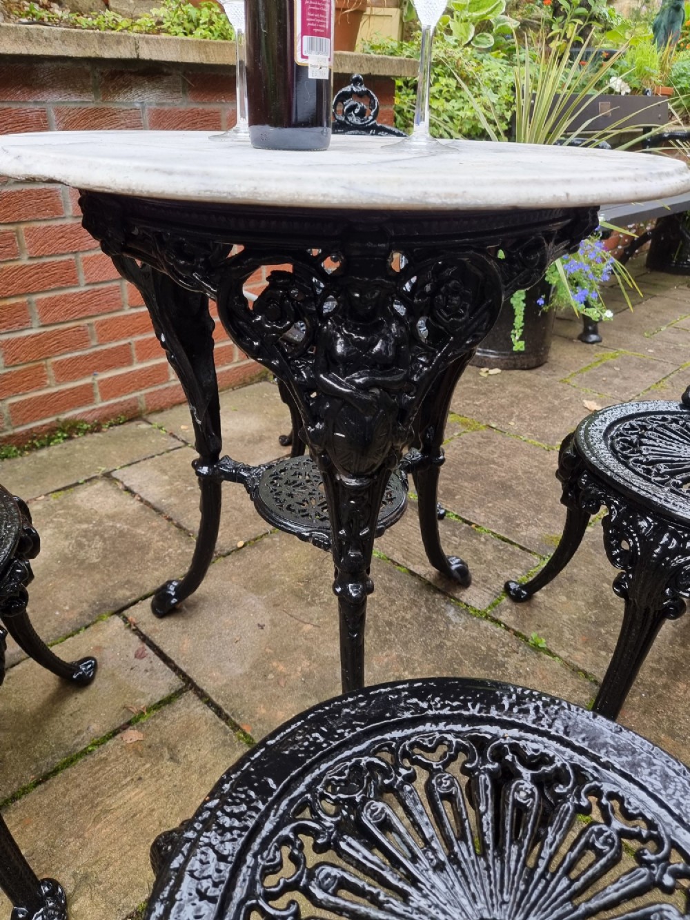 A five-piece garden set featuring a circular white marble-topped table and four black cast iron chairs with ornate pierced floral backrests. View V