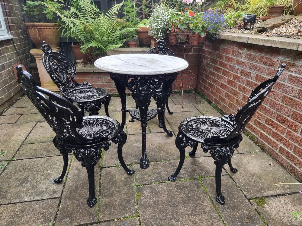 A five-piece garden set featuring a circular white marble-topped table and four black cast iron chairs with ornate pierced floral backrests. View IX