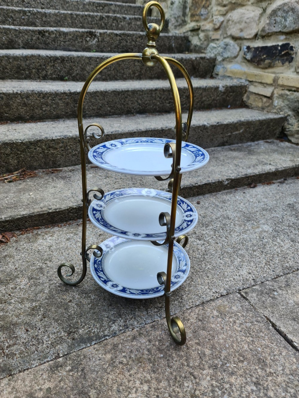A three-tiered Edwardian cake stand featuring a brass metal frame with a top loop handle holding three blue and white ceramic plates. View V