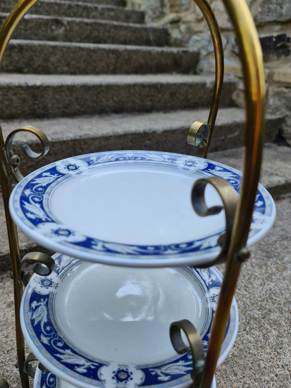 A three-tiered Edwardian cake stand featuring a brass metal frame with a top loop handle holding three blue and white ceramic plates. View VI