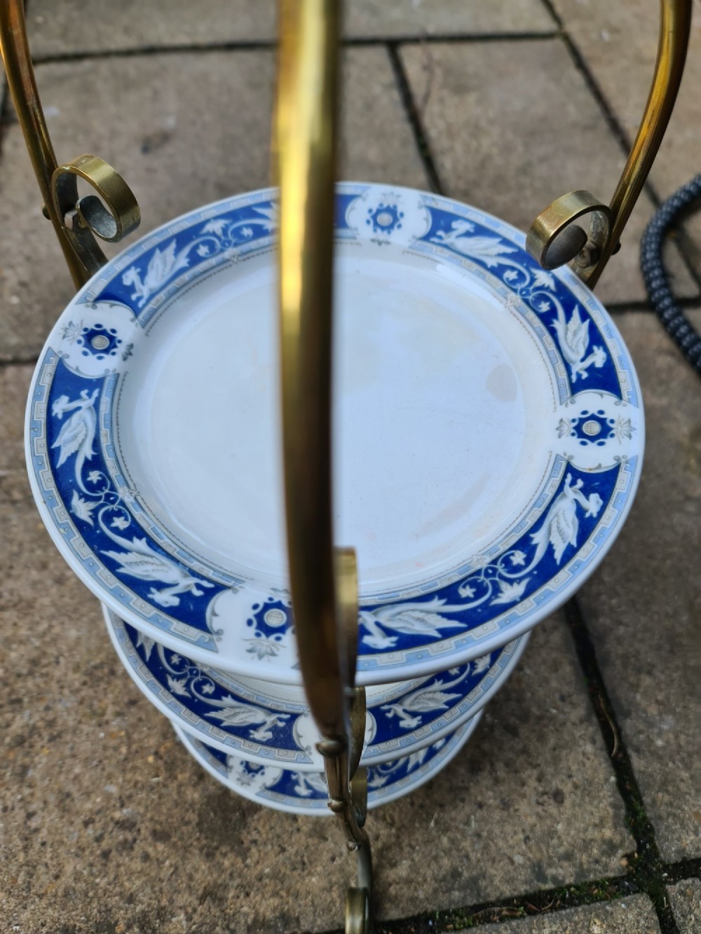 A three-tiered Edwardian cake stand featuring a brass metal frame with a top loop handle holding three blue and white ceramic plates. View VII