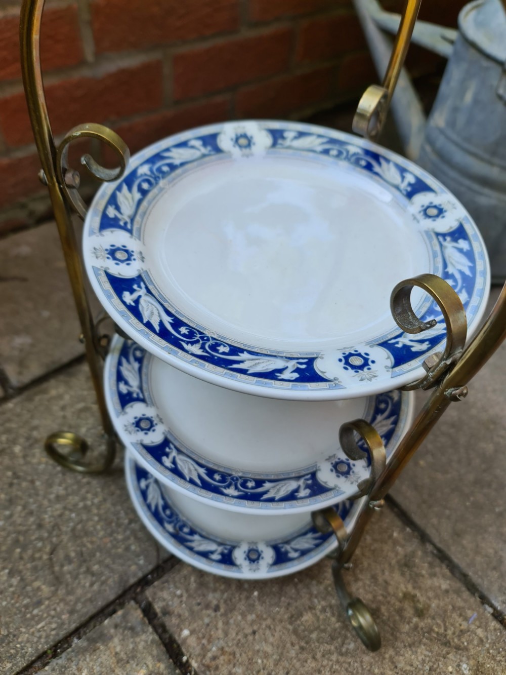 A three-tiered Edwardian cake stand featuring a brass metal frame with a top loop handle holding three blue and white ceramic plates. View VIII
