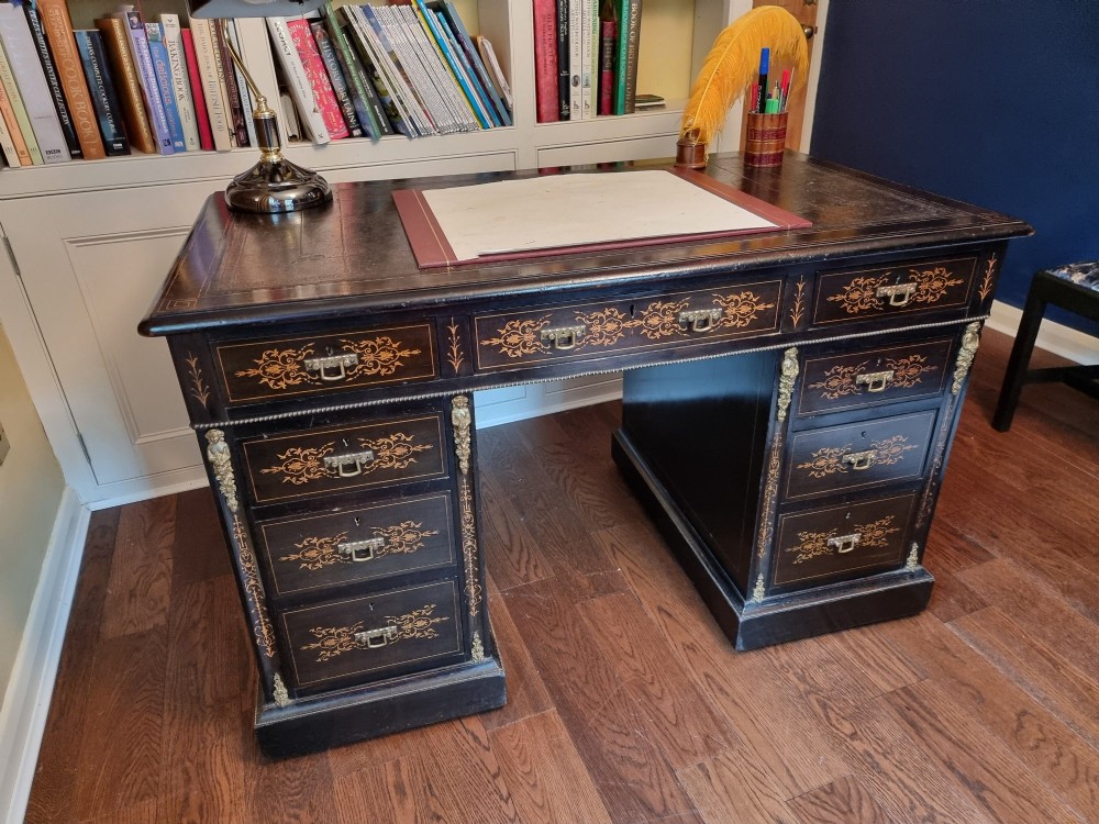 A Victorian ebonised wooden pedestal desk with floral marquetry inlay, leather insert top, brass handles, and gilt corner mounts. View VI