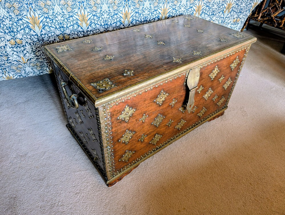 A rectangular padouk wood storage chest decorated with geometric brass mounts, brass studding, side carrying handles, and a central brass lock clasp.
