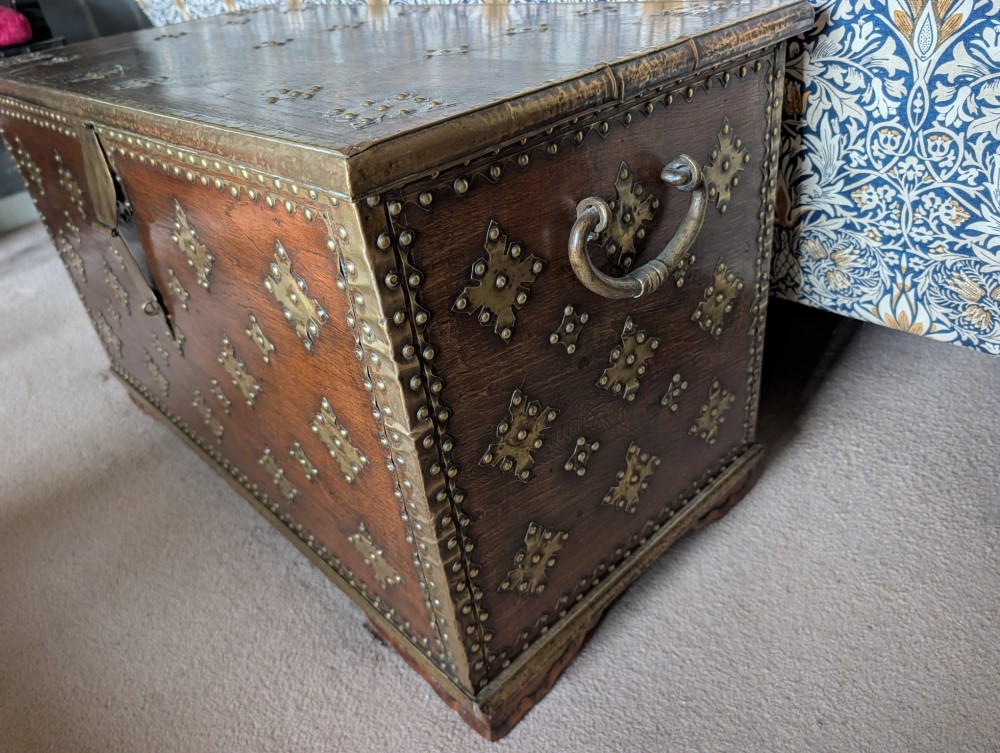 A rectangular padouk wood storage chest decorated with geometric brass mounts, brass studding, side carrying handles, and a central brass lock clasp. View V
