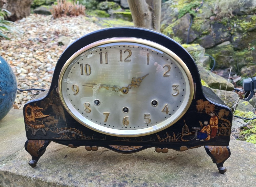 An antique black lacquered mantel clock featuring hand-painted gilt chinoiserie scenes, an oval clock face with gold Arabic numerals, and brass bracket feet. View II