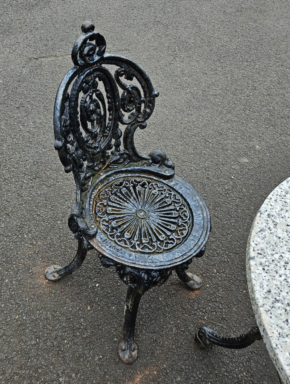 A black cast-iron outdoor bistro set featuring a circular stone-topped table and four matching ornate openwork chairs. View V