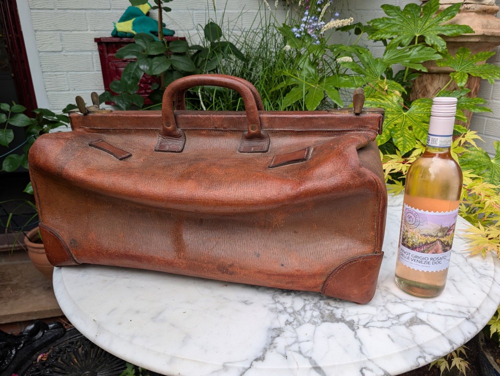 An antique warm brown leather Gladstone travel bag with a brass central lock mechanism, side latches, top carry handle, and stamped monogrammed initials. View IX