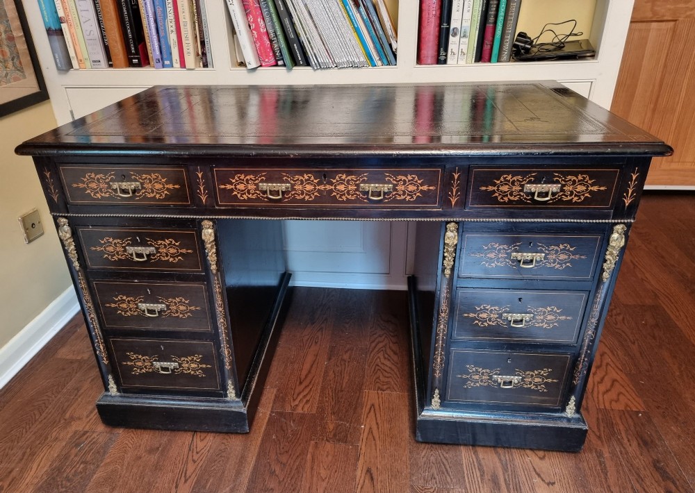 A dark Victorian pedestal desk with marquetry inlay, brass handles, ornate corner mounts, and a leather inset writing surface.