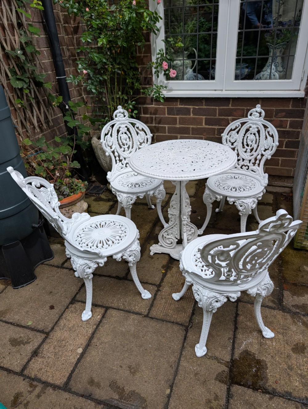 A white painted cast-iron garden set featuring a circular pierced table and four matching ornate chairs with scrollwork backs arranged on a paved stone patio. View III