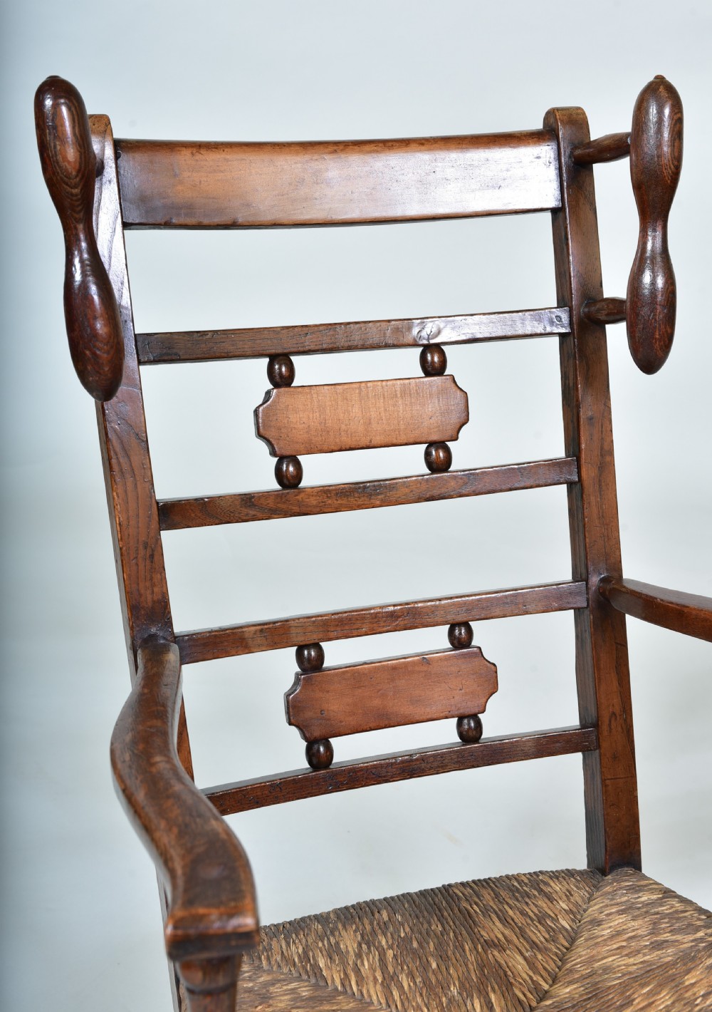 A 19th-century childs dark wood rocking chair featuring a ladder back structure, turned finials, shaped splats, and a woven rush seat. View V
