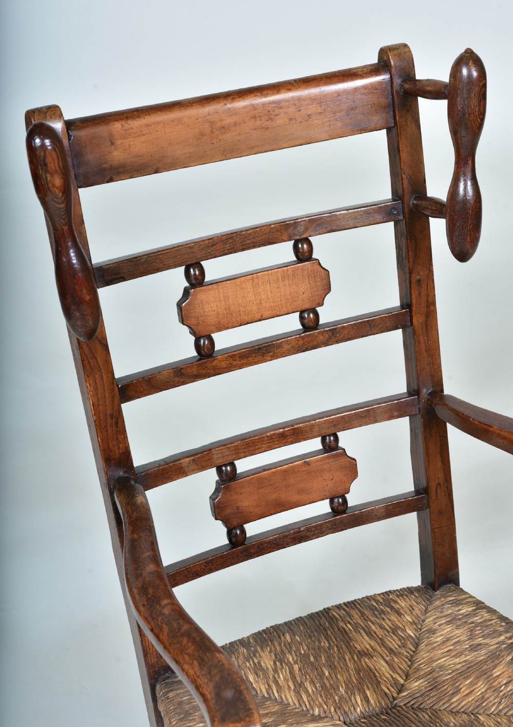 A 19th-century childs dark wood rocking chair featuring a ladder back structure, turned finials, shaped splats, and a woven rush seat. View VI