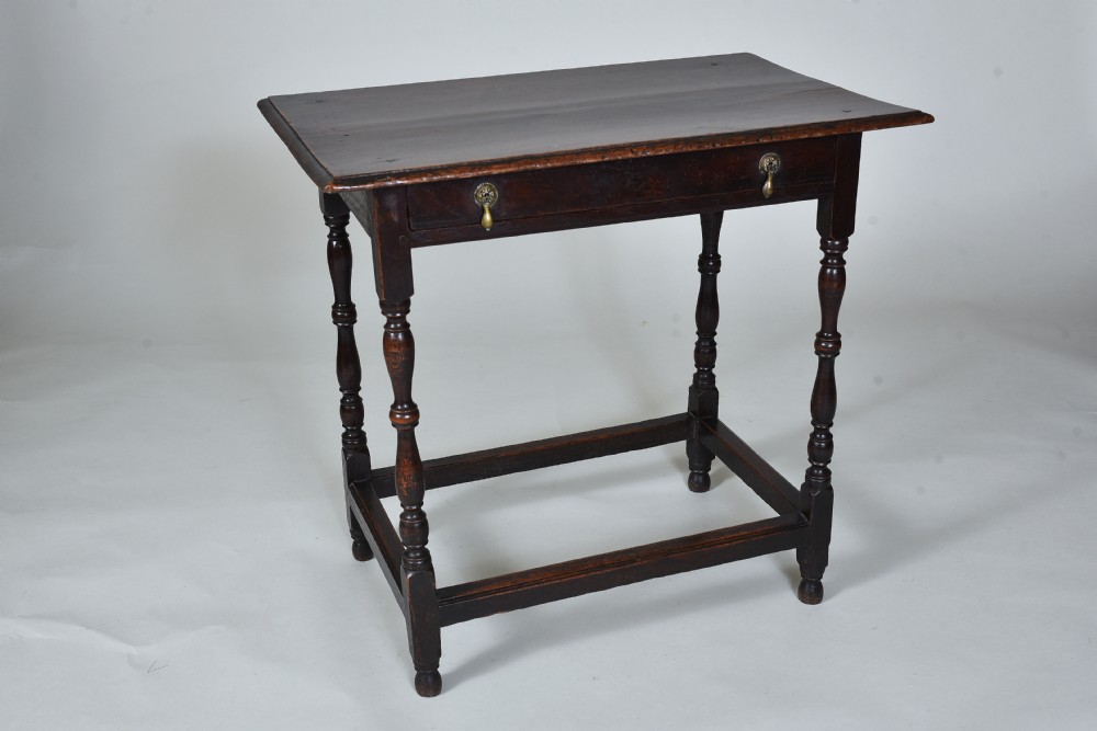 A 17th century dark oak side table featuring a single drawer with brass drop handles, turned legs, and a low box stretcher.