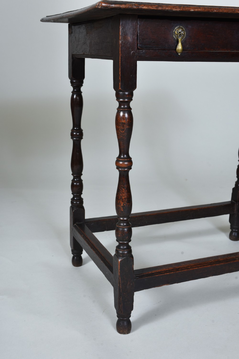 A 17th century dark oak side table featuring a single drawer with brass drop handles, turned legs, and a low box stretcher. View V