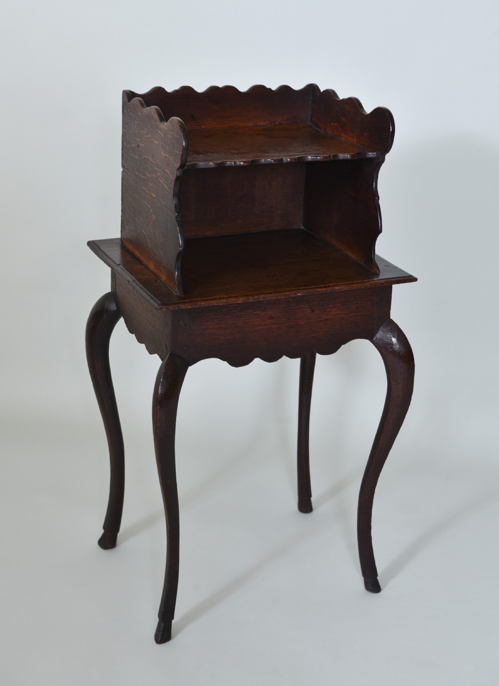 An 18th-century dark oak side table featuring curved cabriole legs, a scalloped apron, and a raised two-tier scalloped gallery shelf.