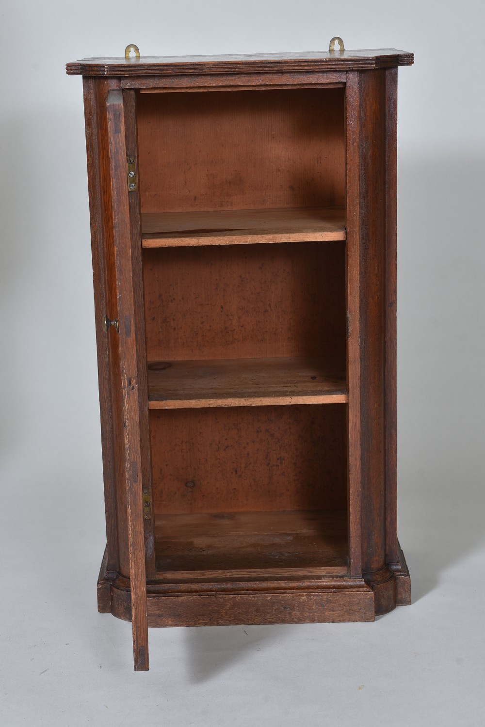 A narrow 19th-century dark brown oak wall cupboard with a three-panel door, brass knob, and metal hanging brass loops at the top. View II