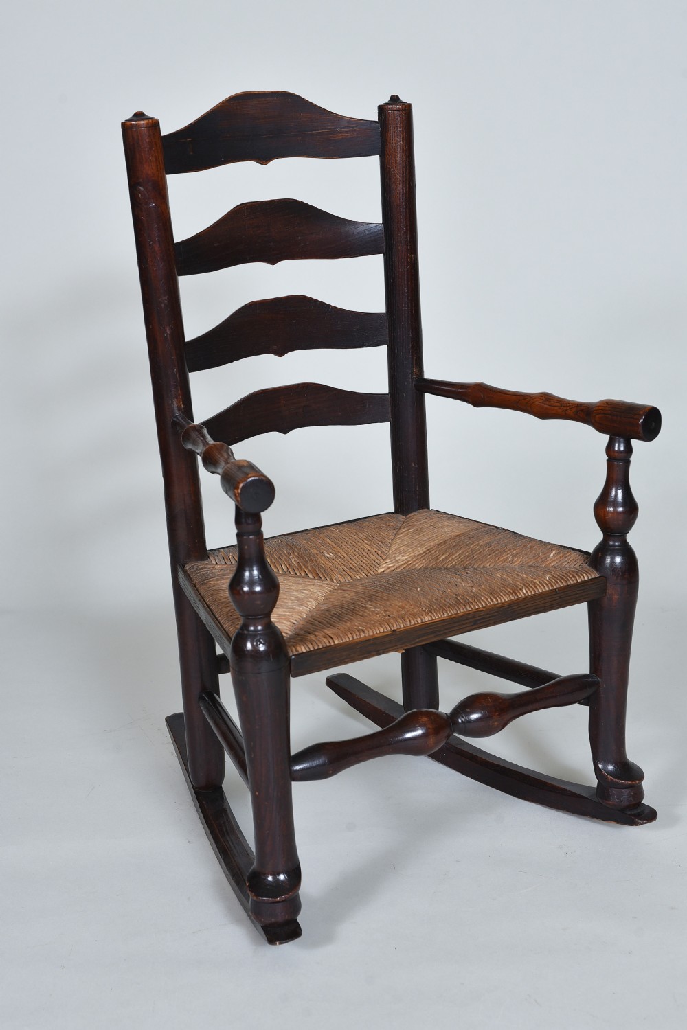 An early nineteenth-century childs rocking chair crafted from dark brown wood with a shaped ladderback backrest, turned armrests, and a woven rush seat.