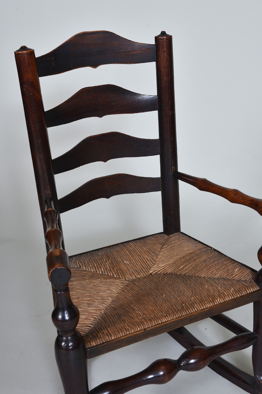 An early nineteenth-century childs rocking chair crafted from dark brown wood with a shaped ladderback backrest, turned armrests, and a woven rush seat. View II
