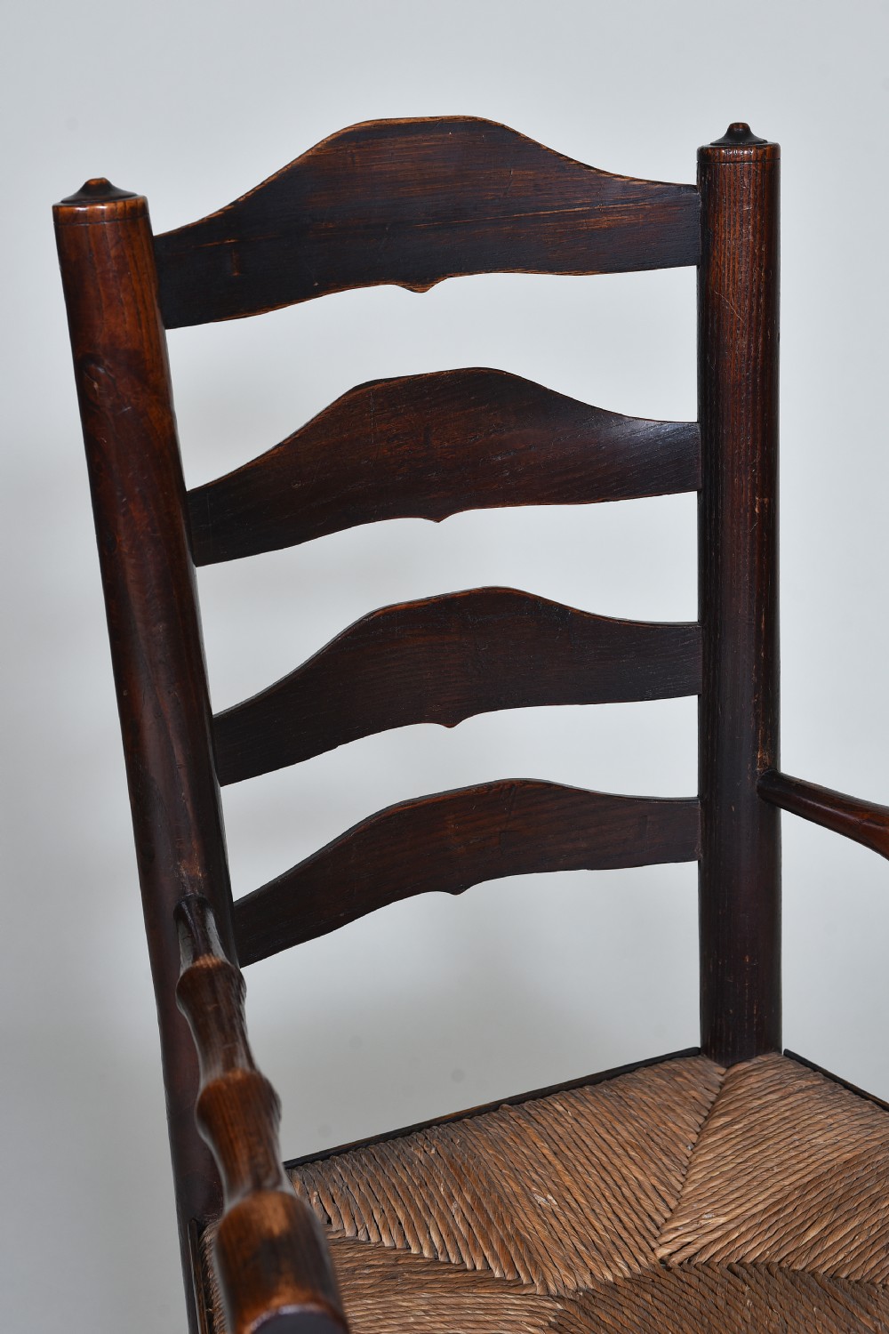 An early nineteenth-century childs rocking chair crafted from dark brown wood with a shaped ladderback backrest, turned armrests, and a woven rush seat. View III