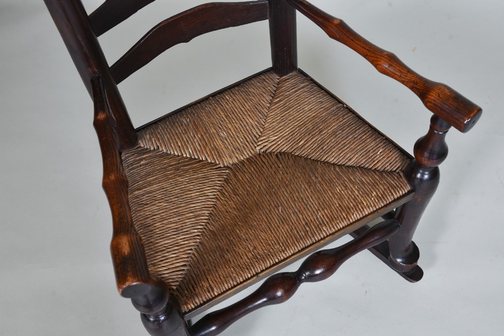 An early nineteenth-century childs rocking chair crafted from dark brown wood with a shaped ladderback backrest, turned armrests, and a woven rush seat. View V