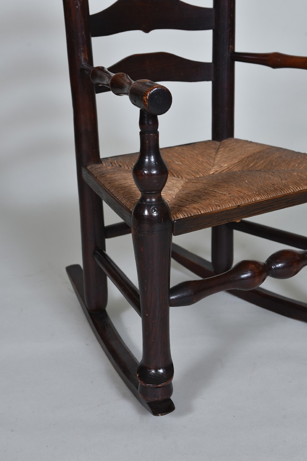 An early nineteenth-century childs rocking chair crafted from dark brown wood with a shaped ladderback backrest, turned armrests, and a woven rush seat. View VI