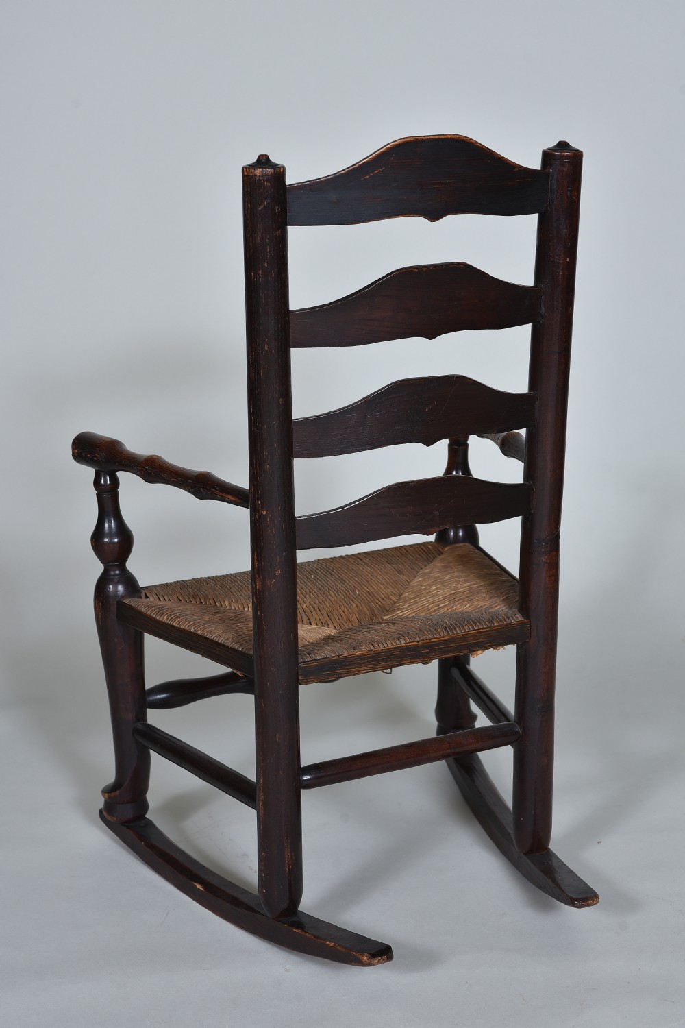 An early nineteenth-century childs rocking chair crafted from dark brown wood with a shaped ladderback backrest, turned armrests, and a woven rush seat. View VIII