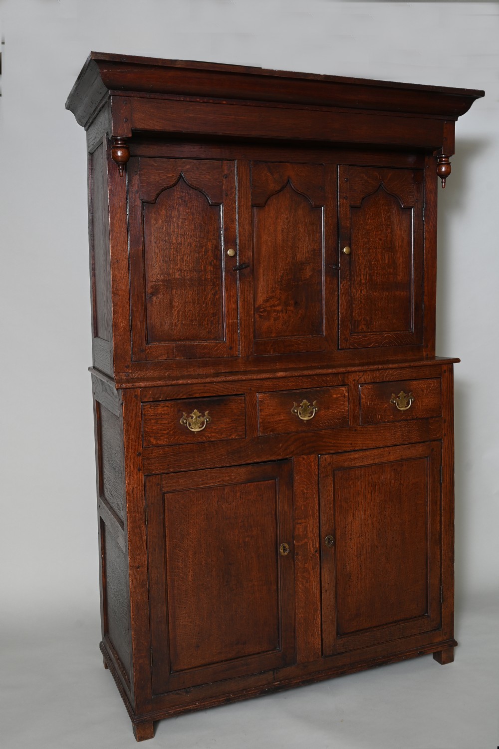 An 18th-century dark oak Welsh press cupboard featuring upper ogee arched panel doors, turned drop finials, three middle drawers, and a double-door base.