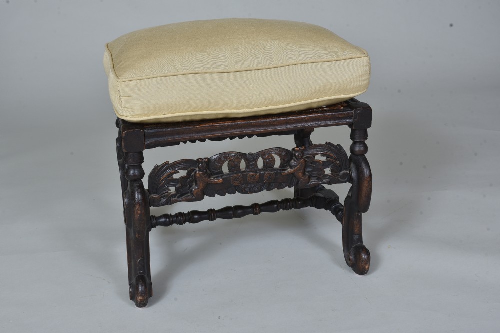 An antique dark brown carved wooden stool featuring scroll legs, an ornate carved apron, and a cream upholstered cushion.