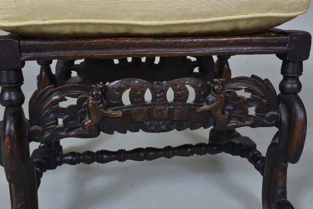An antique dark brown carved wooden stool featuring scroll legs, an ornate carved apron, and a cream upholstered cushion. View II